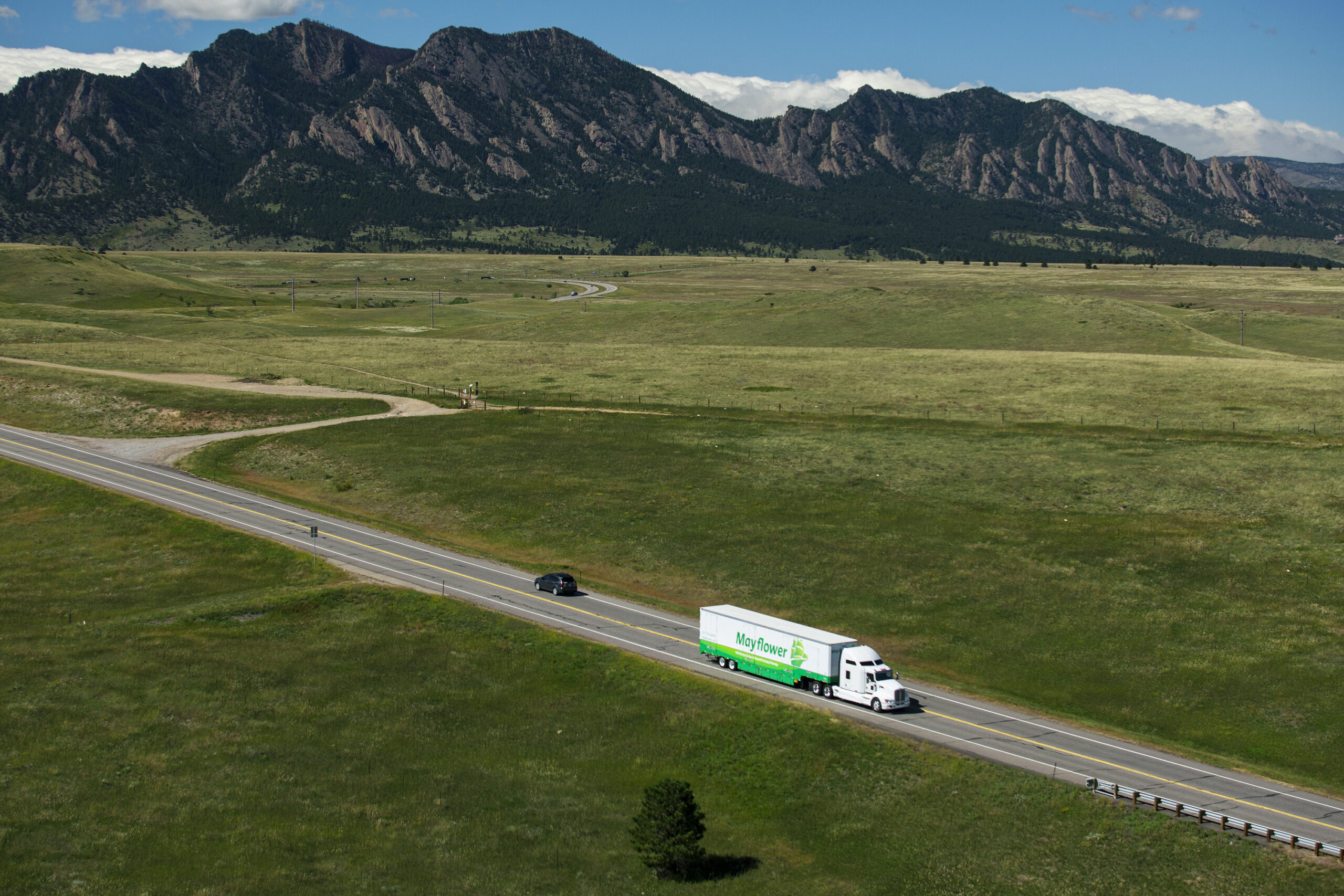 Wide landscape of grassy fields with a winding road and a white-green Mayflower truck driving past, mountains in the distance under a blue sky.