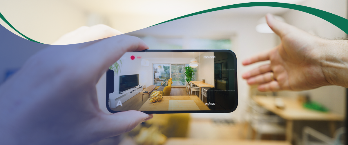 Person records a living room interior on a smartphone screen, showing a couch, coffee table, plants, and large windows in a sunny open space.