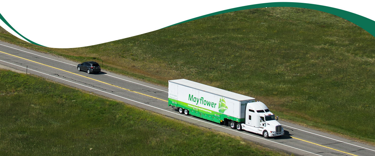 White semi-truck with a green Mayflower trailer driving on a two-lane highway with a small car ahead on the left side of the road, grassy hills surrounding.