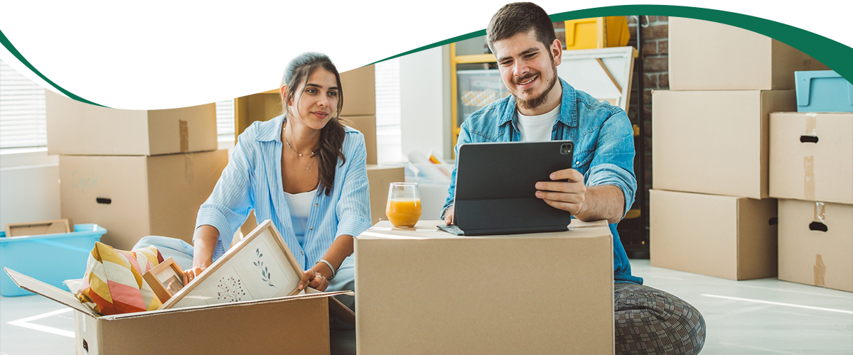 Couple packing and organizing in a sunny room, using a tablet while surrounded by moving boxes and supplies