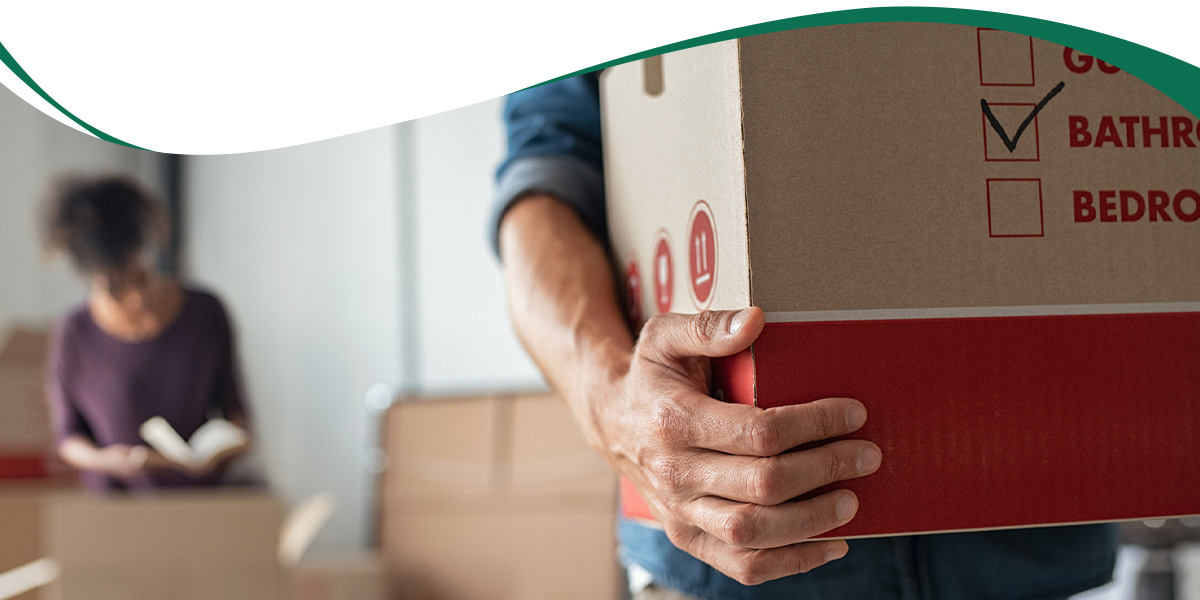 Person in foreground carrying a large cardboard box during a move, with a woman reading in the background amid more boxes.