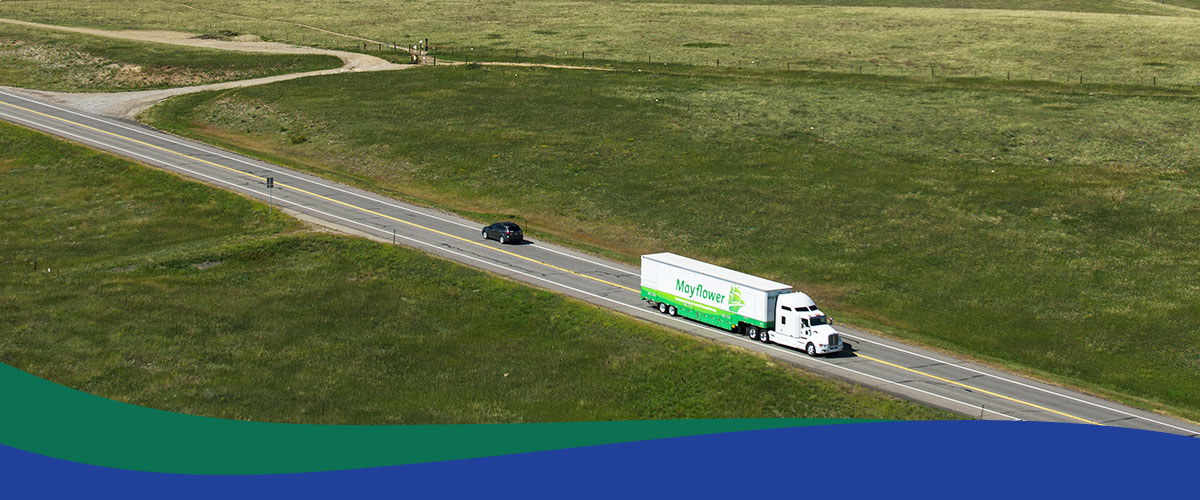 Aerial view of a white Mayflower moving truck on a rural two-lane highway with a dark car behind, surrounded by green fields.