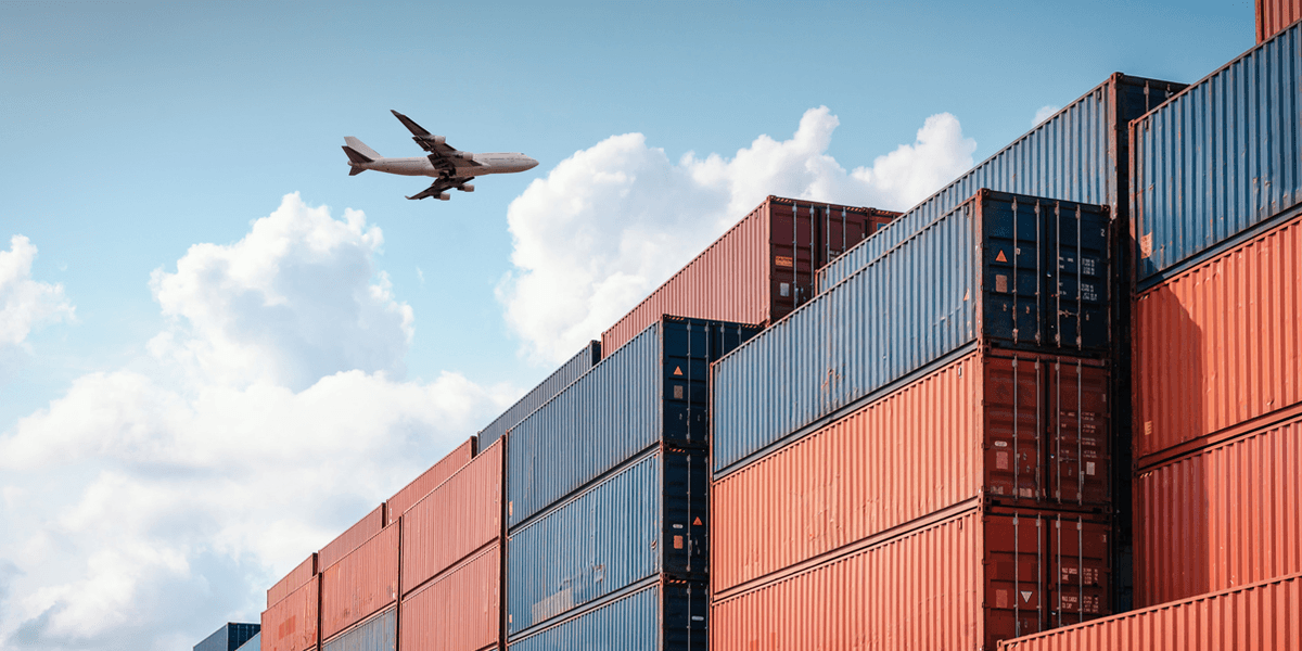 Stacked red and blue shipping containers under a blue sky with an airplane flying overhead