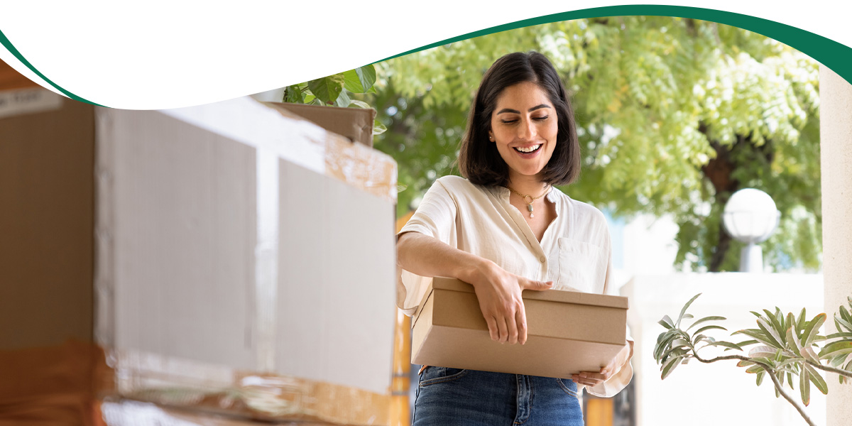 Woman smiling while holding a cardboard box outdoors, with more packages nearby.