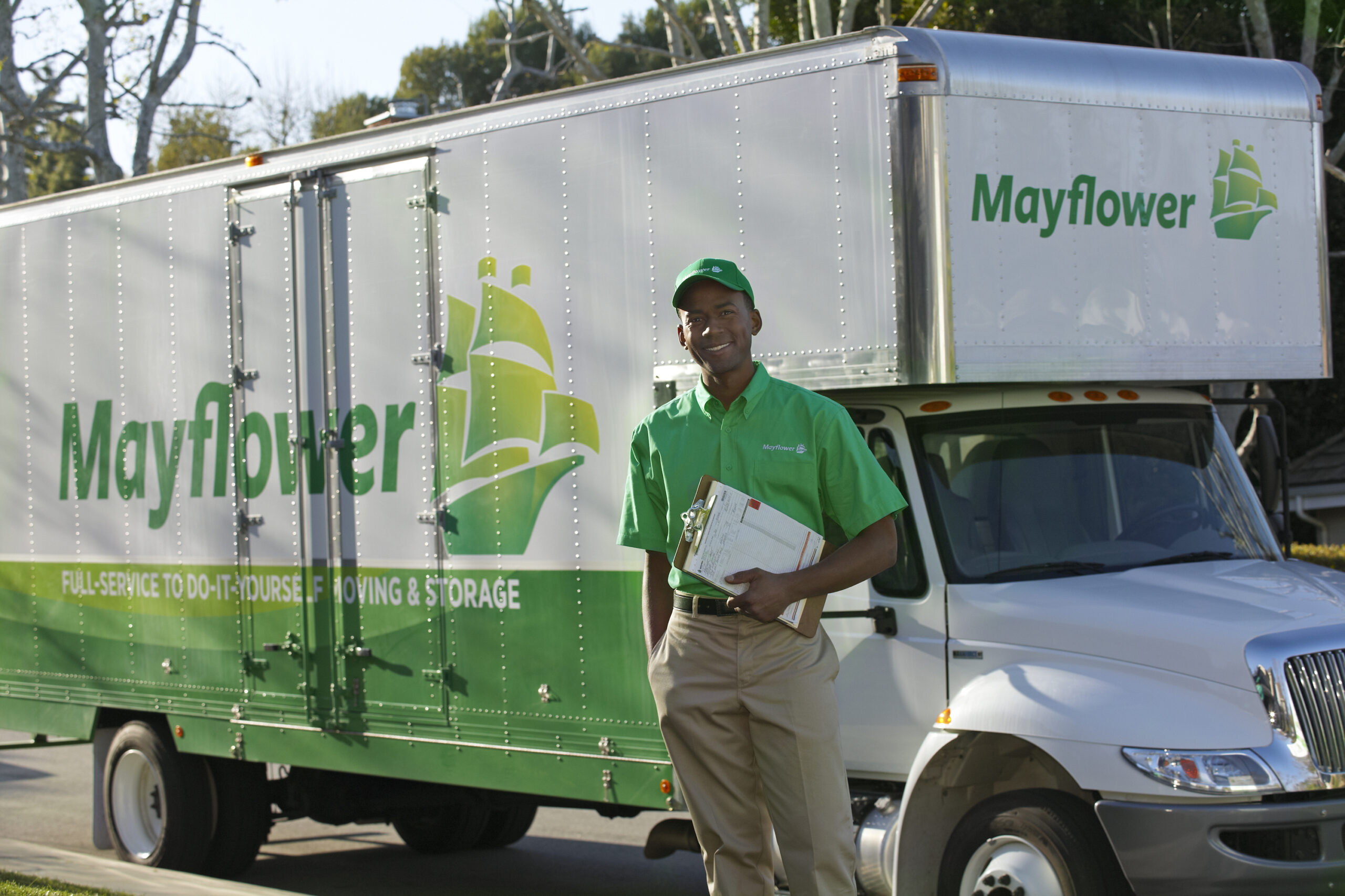 Delivery worker in a green uniform standing beside a large Mayflower moving truck