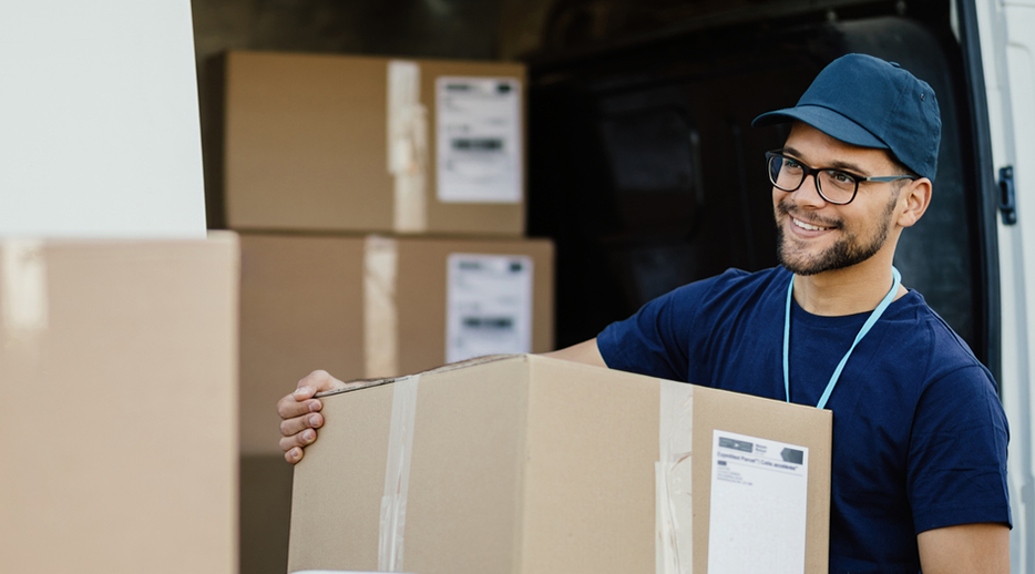 Delivery worker in a blue shirt and cap lifts a large taped cardboard box near an open van.